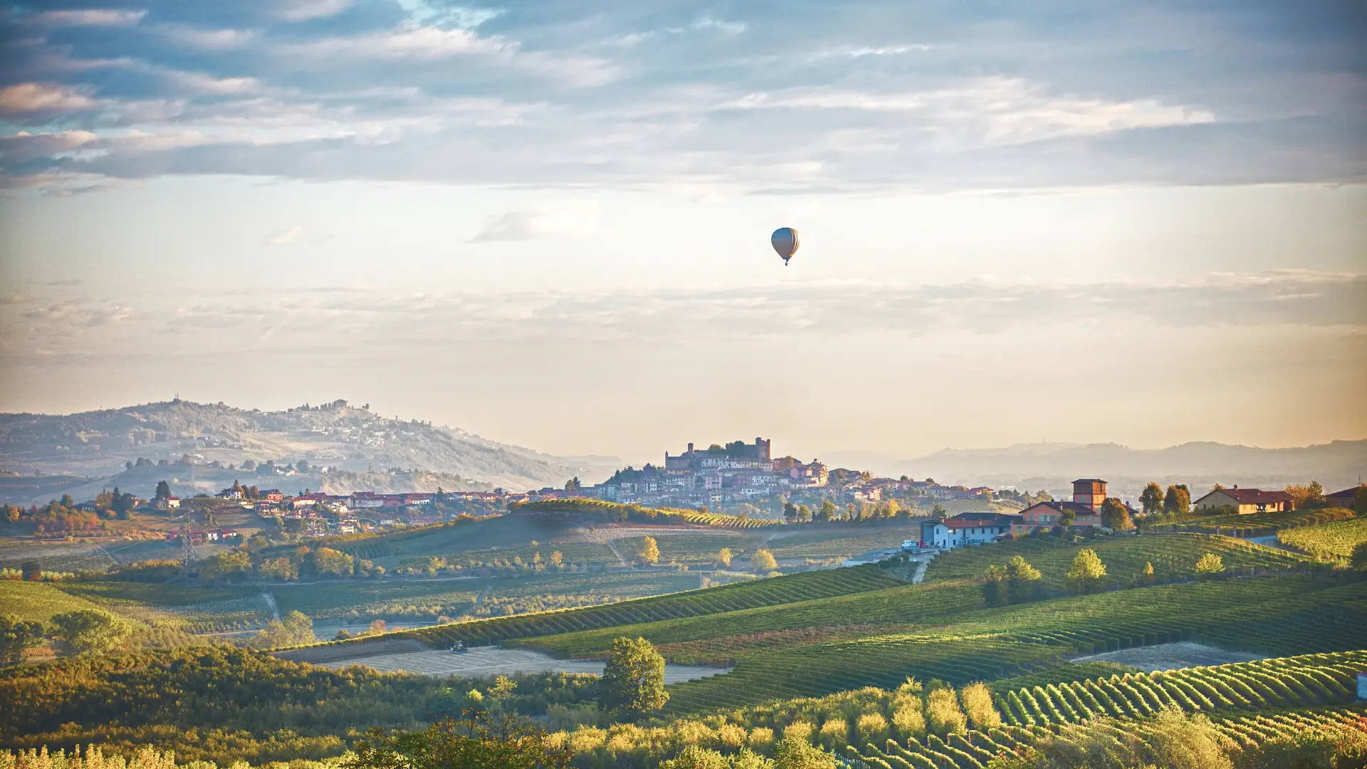 A vibrant hot air balloon soars gently above the verdant hills and vineyards of Langhe-Roero-Monferrato. The golden sunlight and lush, green fields create a tranquil atmosphere, capturing the serene beauty of springtime in northern Italy, perfect for wine lovers and nature enthusiasts