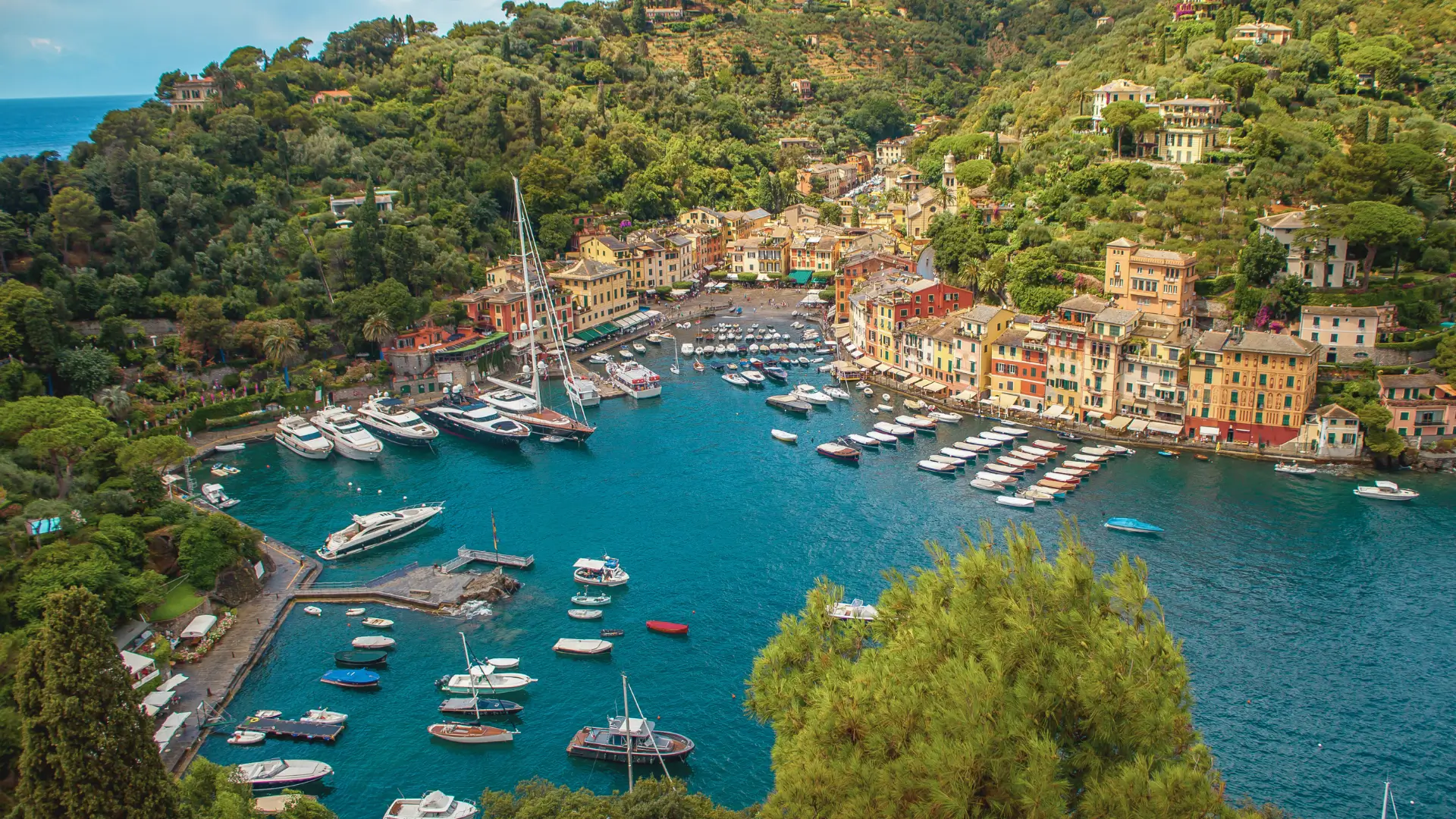 The picturesque Portofino harbour features pastel-hued houses lining the waterfront, overlooking sparkling, turquoise waters. The warm, clear light highlights the quiet charm of this iconic Ligurian coastal village