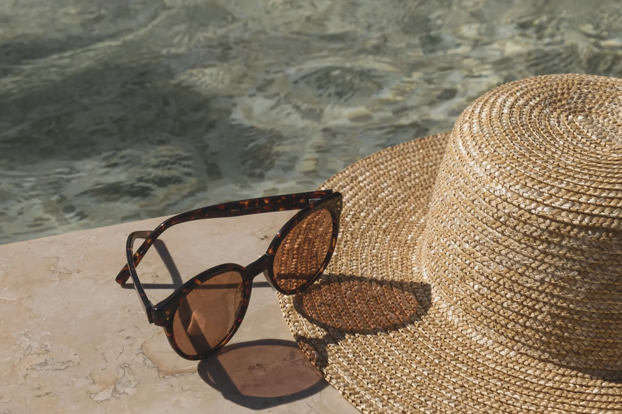 A sun hat and sunglasses beside a sparkling pool on a warm afternoon.